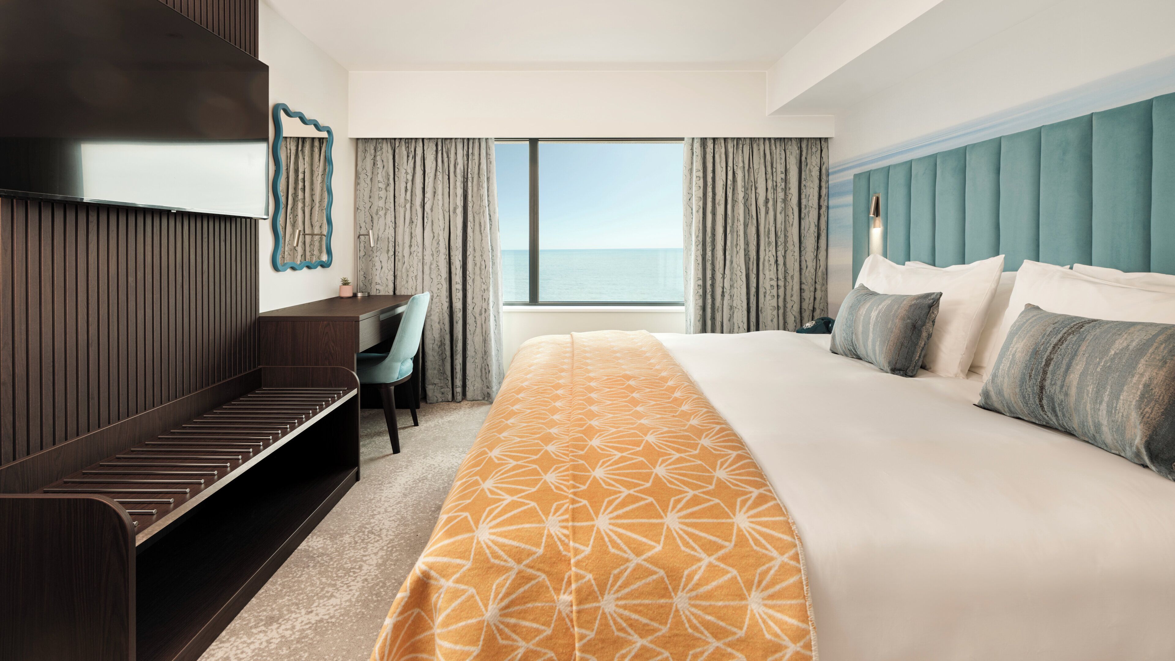 A hotel room with a large bed, a desk, a chair, a TV, and a view of the sea.