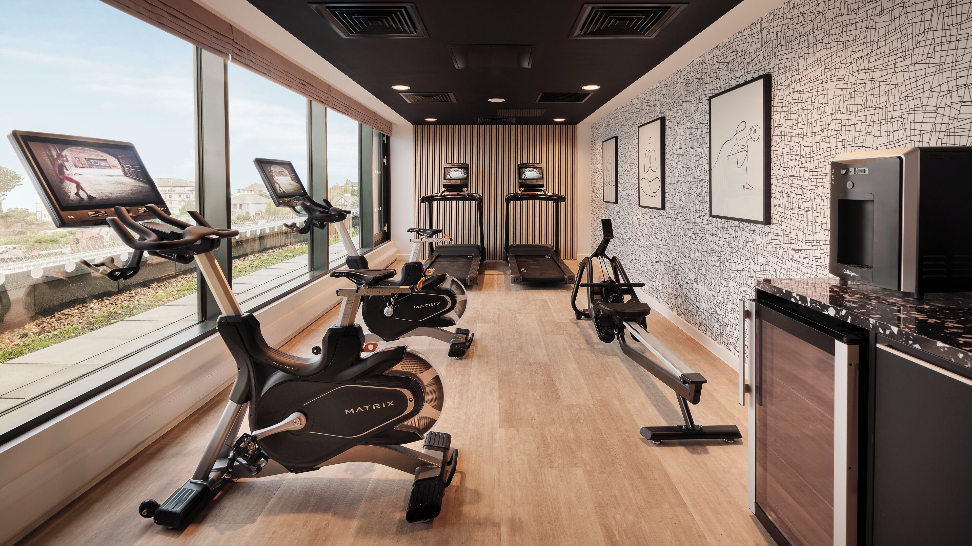 A modern gym with stationary bikes, treadmills, and a rowing machine. It features large windows, a patterned wall, and a kitchen area.
