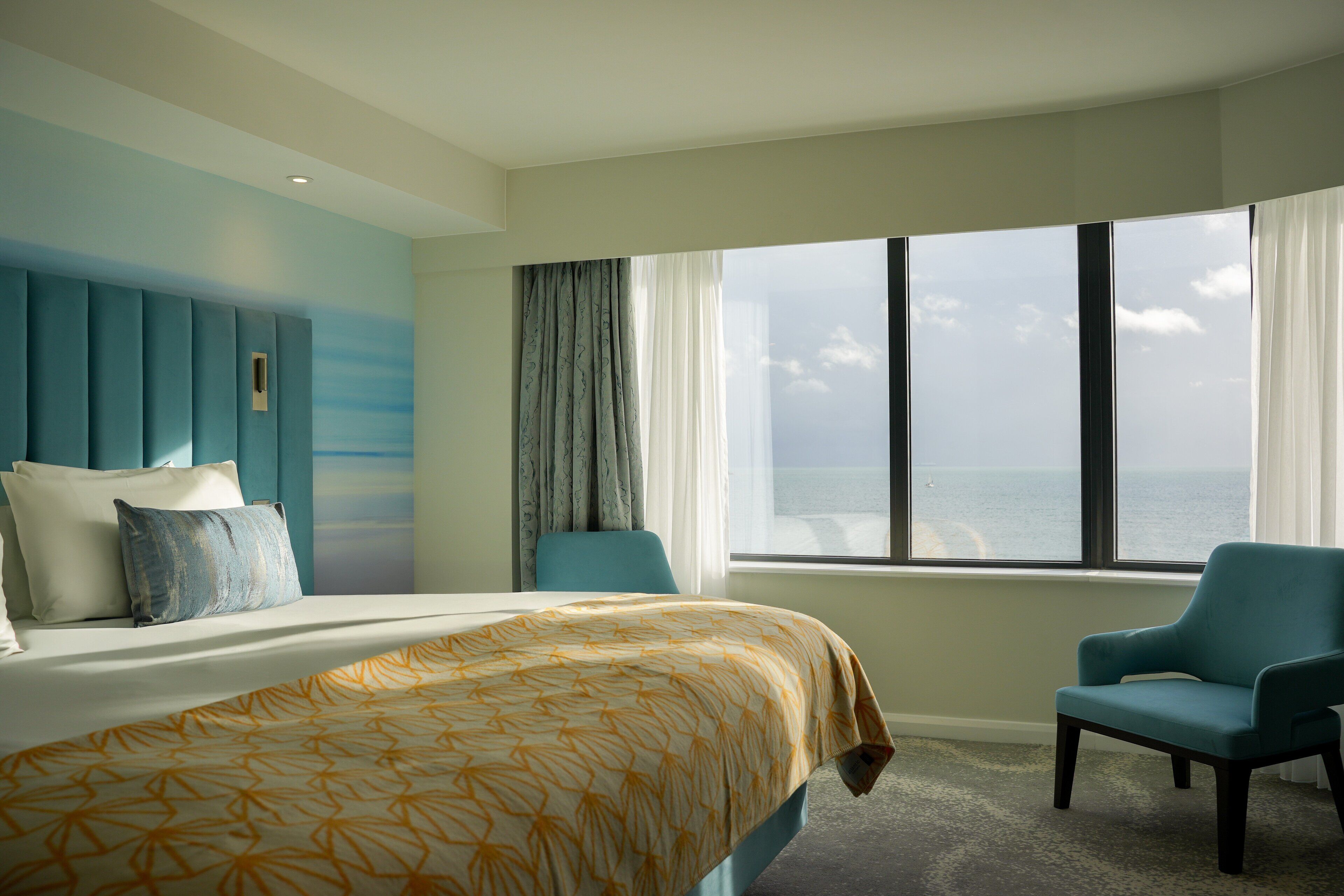 Une chambre d’hôtel avec un grand lit, une vue sur l’océan, une fenêtre avec des rideaux et un fauteuil turquoise.