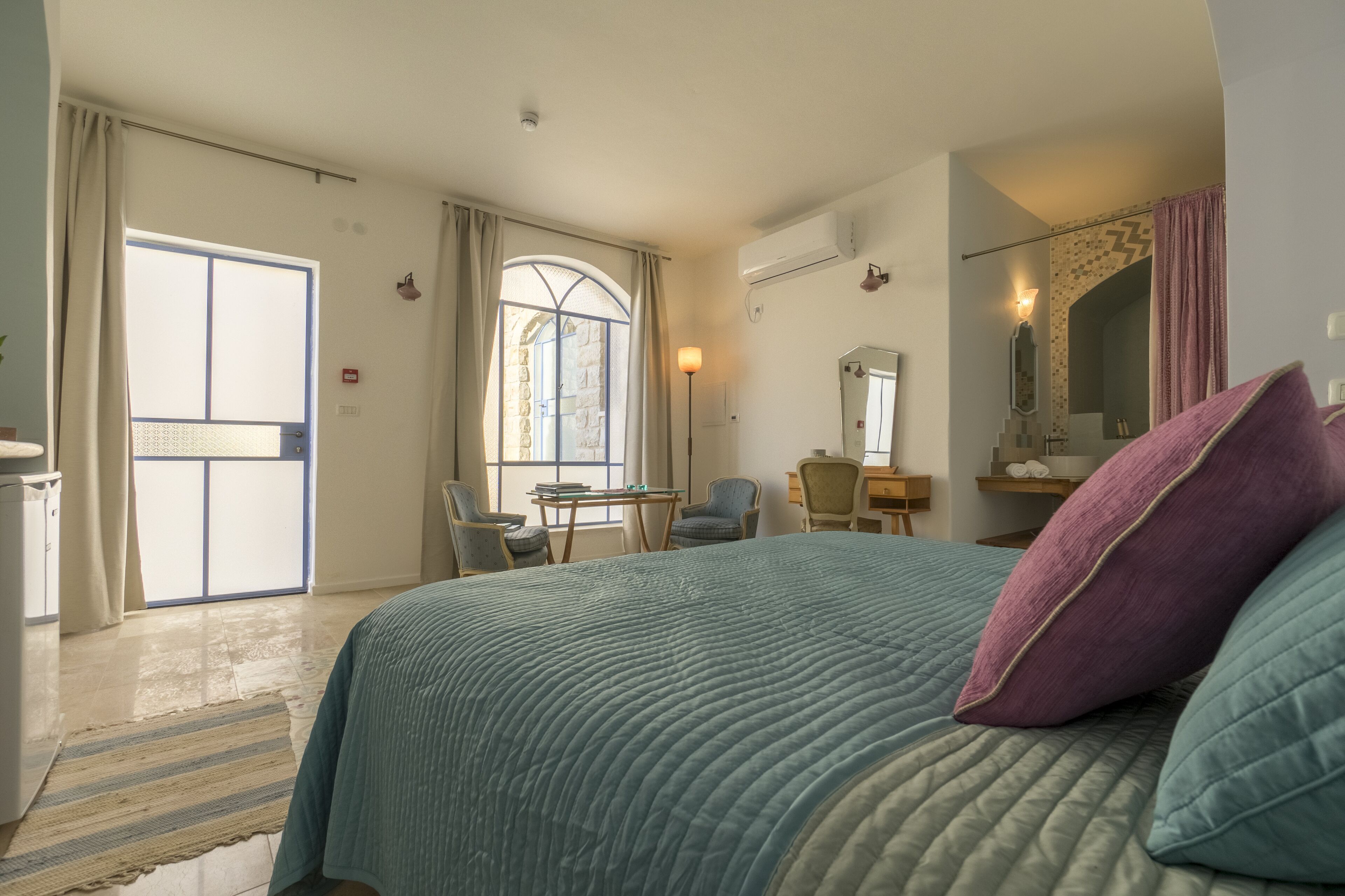 A bedroom with a large bed, a desk, and a chair. There is a window with curtains, a mirror, and a door leading to a balcony.