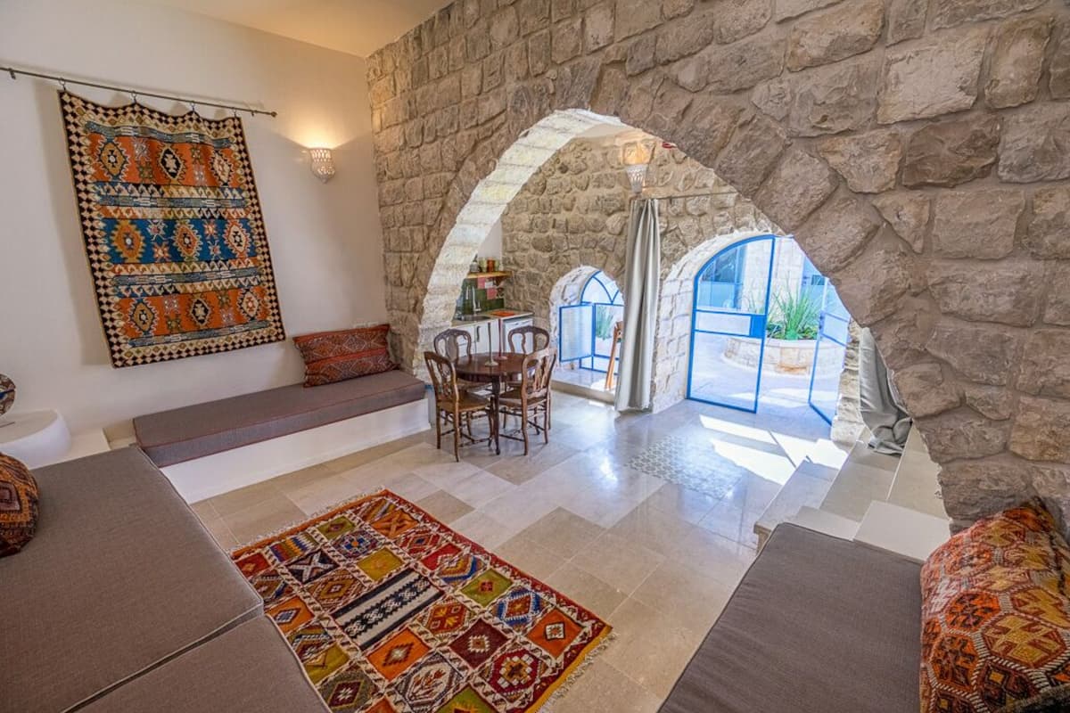 A cozy living room with a stone wall, a large colorful rug, and a seating area with a view through an archway to a bright room.