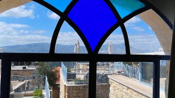 A view through a stained glass window with a geometric design, overlooking a cityscape with buildings, a stone wall, and a metal fence.