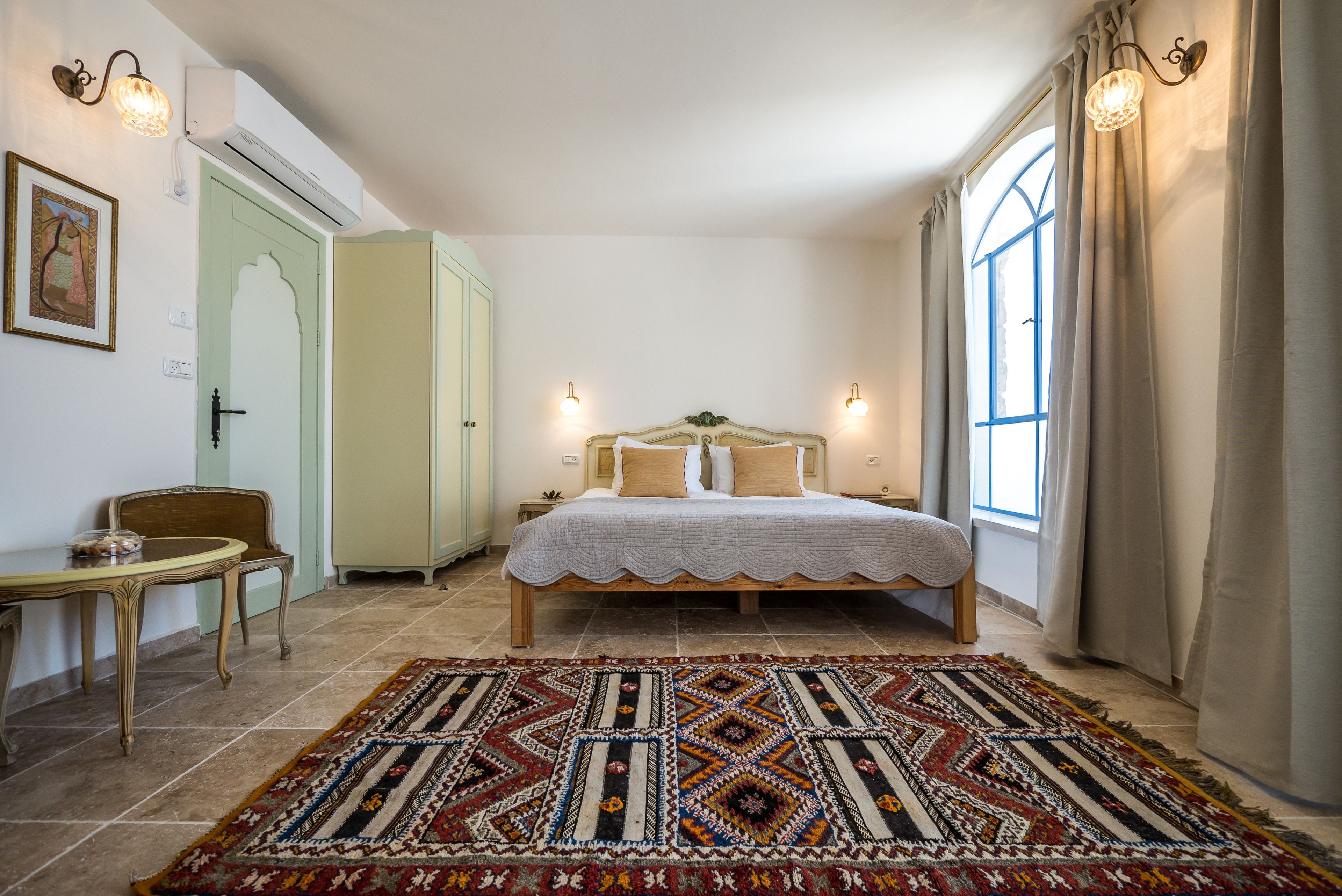 Un dormitorio con una cama, una mesita de noche, una silla, una mesa y un amplio ventanal con cortinas.