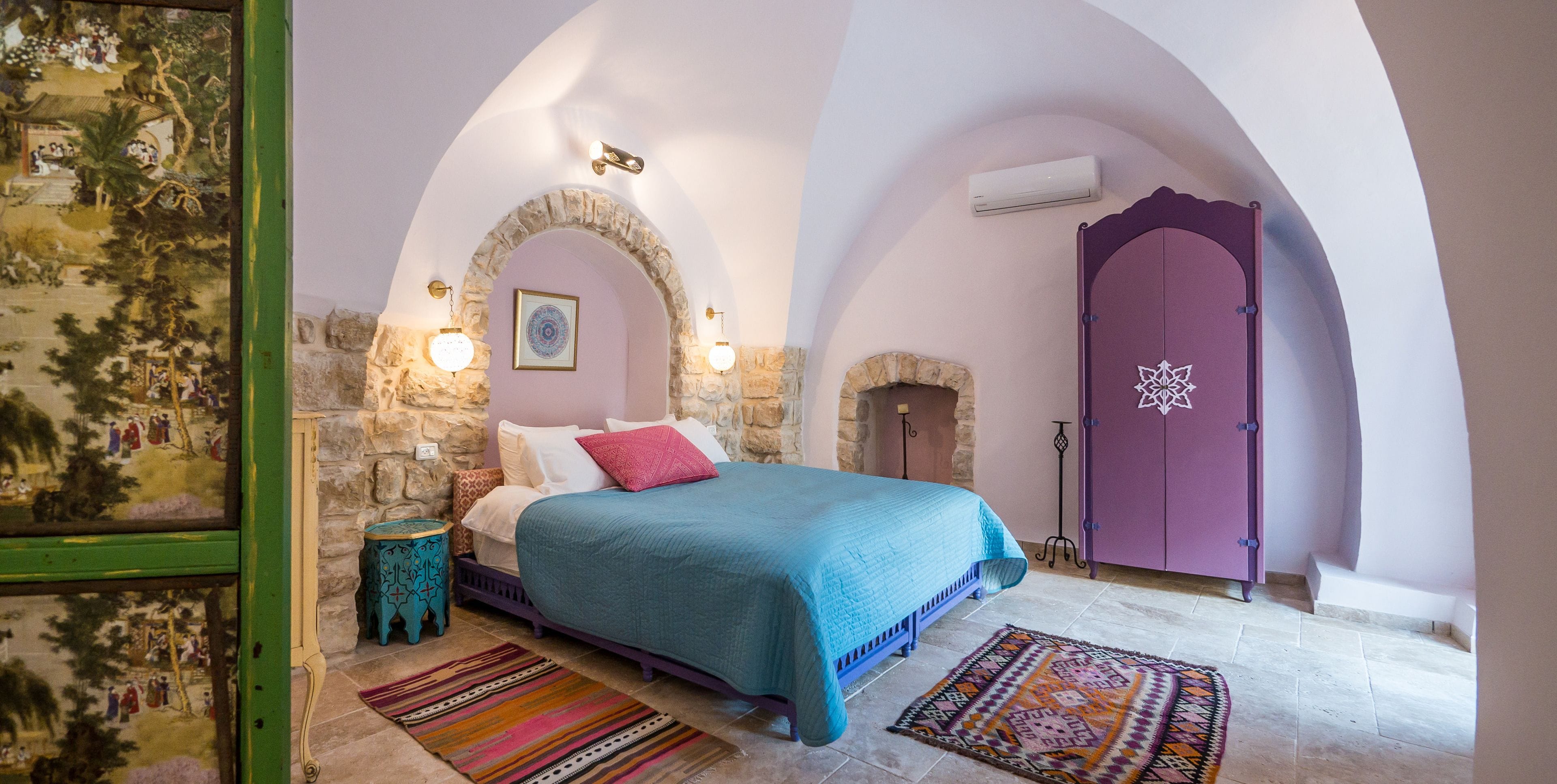 Une chambre à coucher avec un lit, une armoire violette, un mur en pierre et une fresque murale.