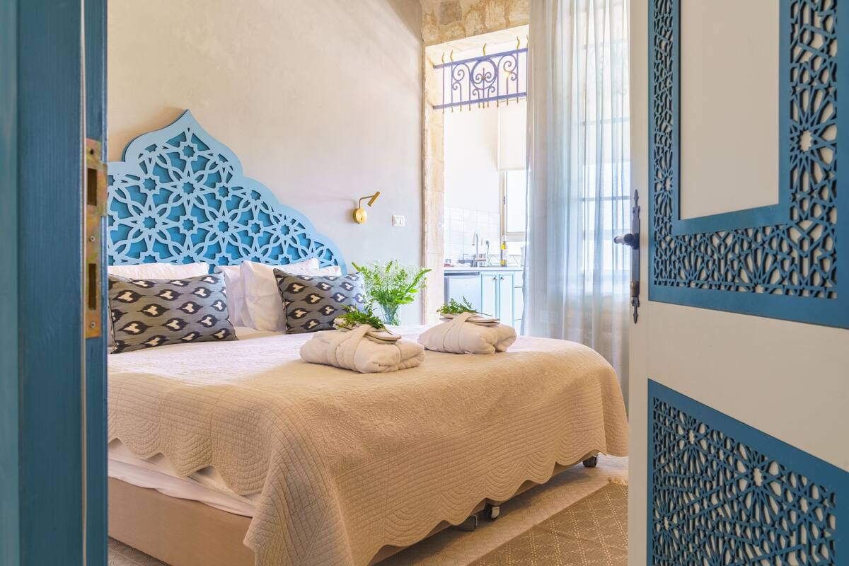 A neatly made bed with a patterned headboard, a bedside table with a lamp, and a window with a decorative metal grille.