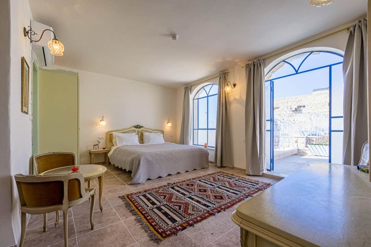 A bedroom with a bed, bedside tables, a chair, a small table, a patterned rug, and large windows with curtains.
