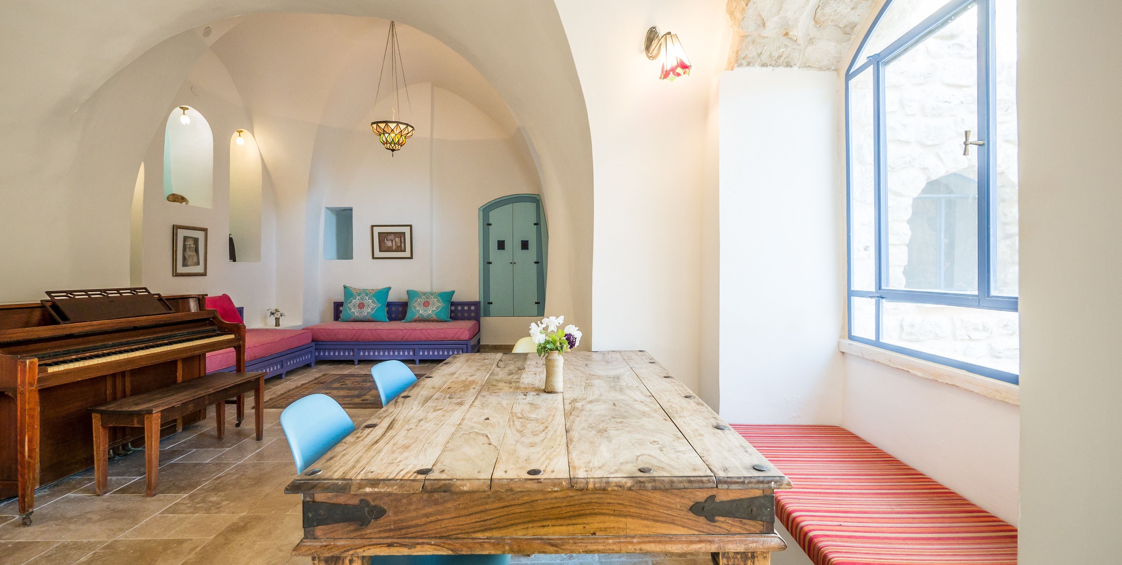 A room with a wooden table, blue chairs, a piano, and a cozy seating area with cushions.