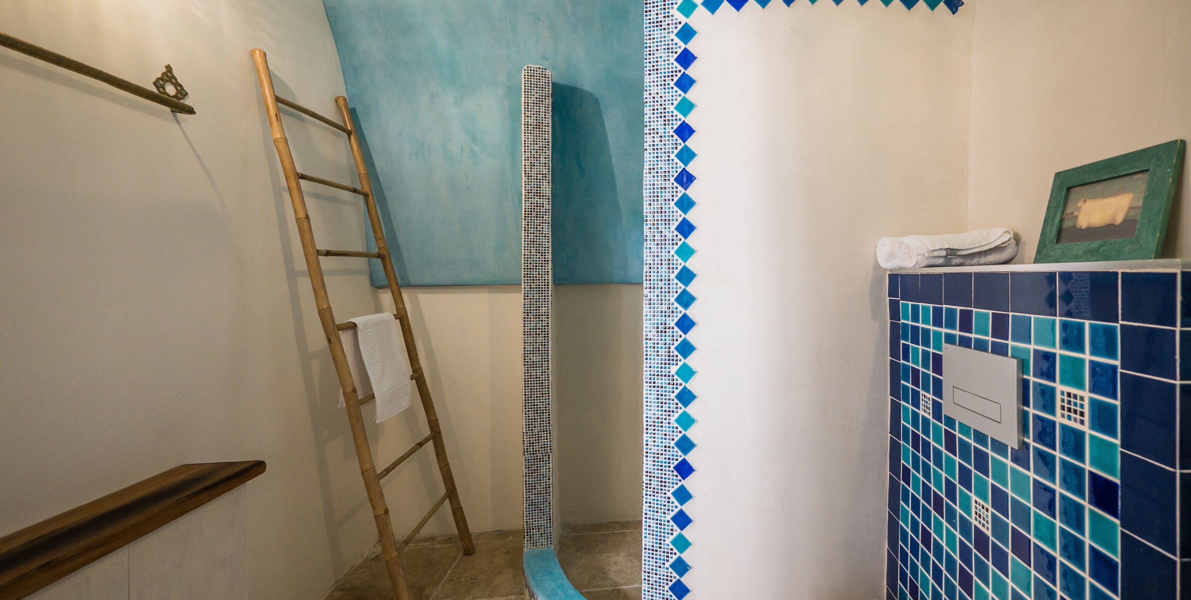 Un baño con una escalera de madera apoyada en la pared, una zona de ducha con azulejos azules y blancos, y un perchero con una toalla blanca.