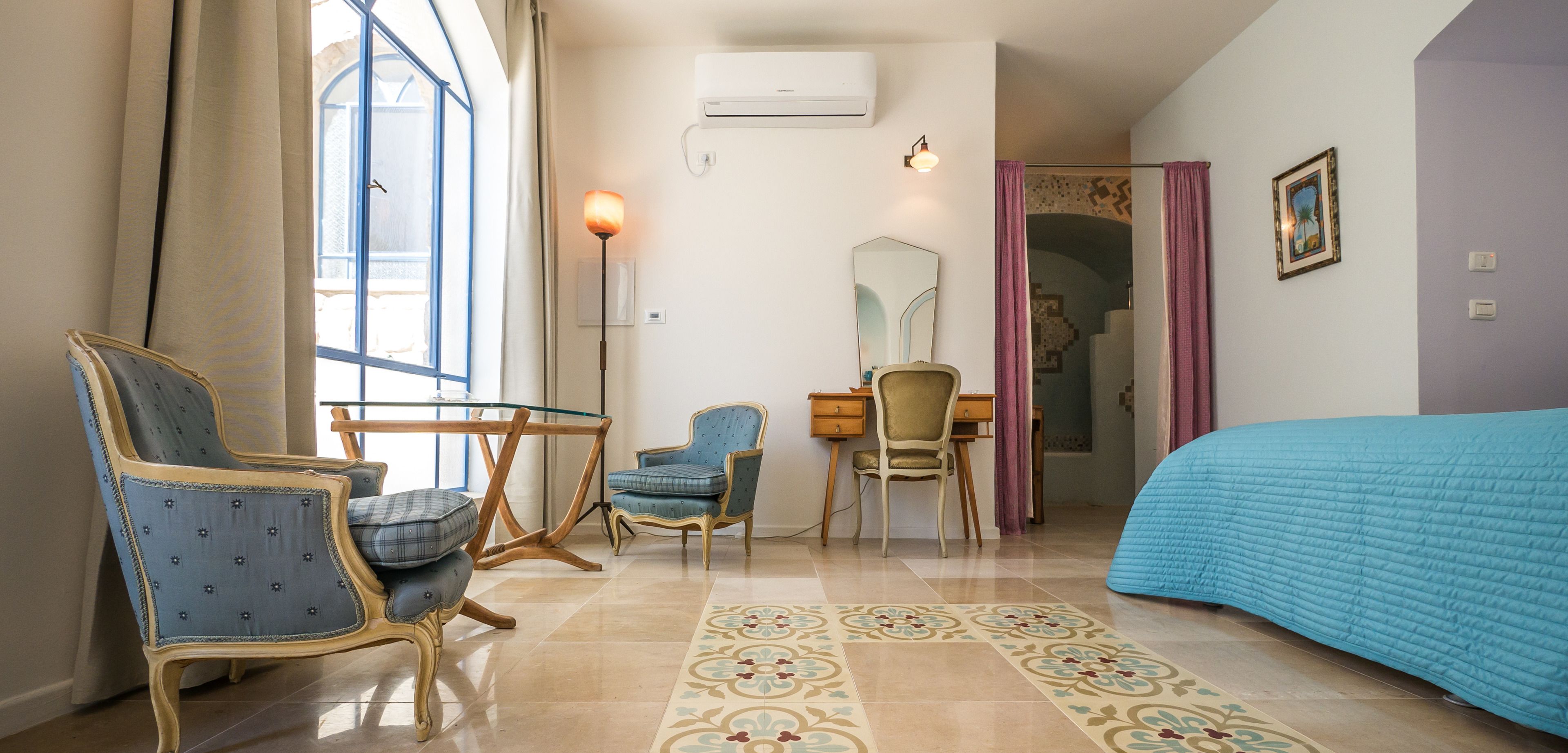 Une chambre d’hôtel comprenant un lit, un bureau avec une chaise, une chaise avec un coussin, une lampe et une fenêtre avec des rideaux.