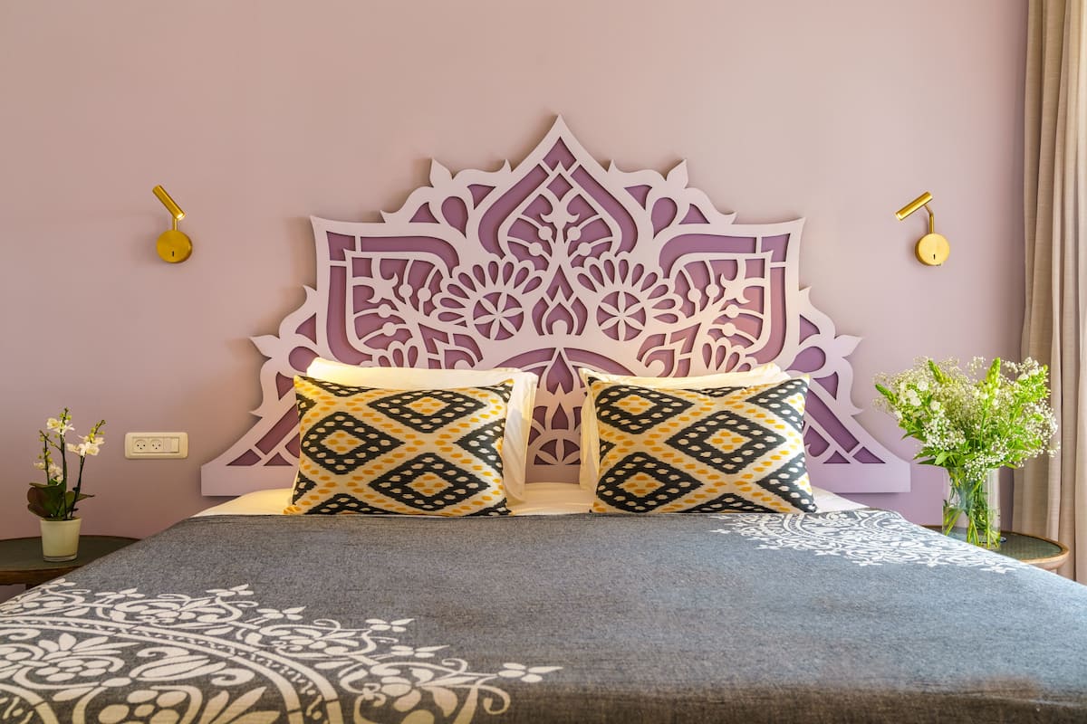 A bed with a decorative headboard, two patterned pillows, a vase with flowers, and a wall socket.