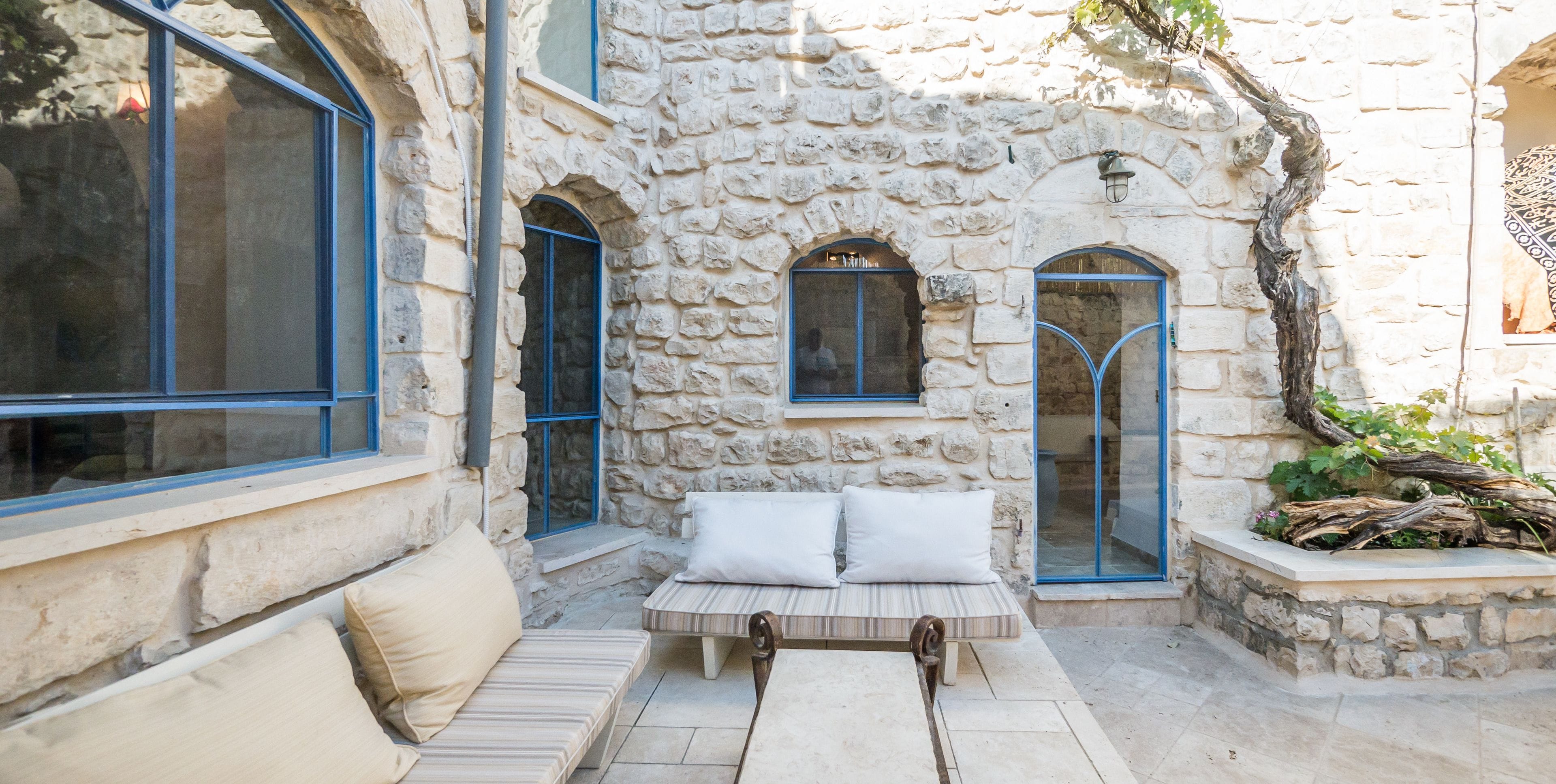 A stone-walled outdoor seating area with a bench and cushions, a small table, and a window with blue frames.
