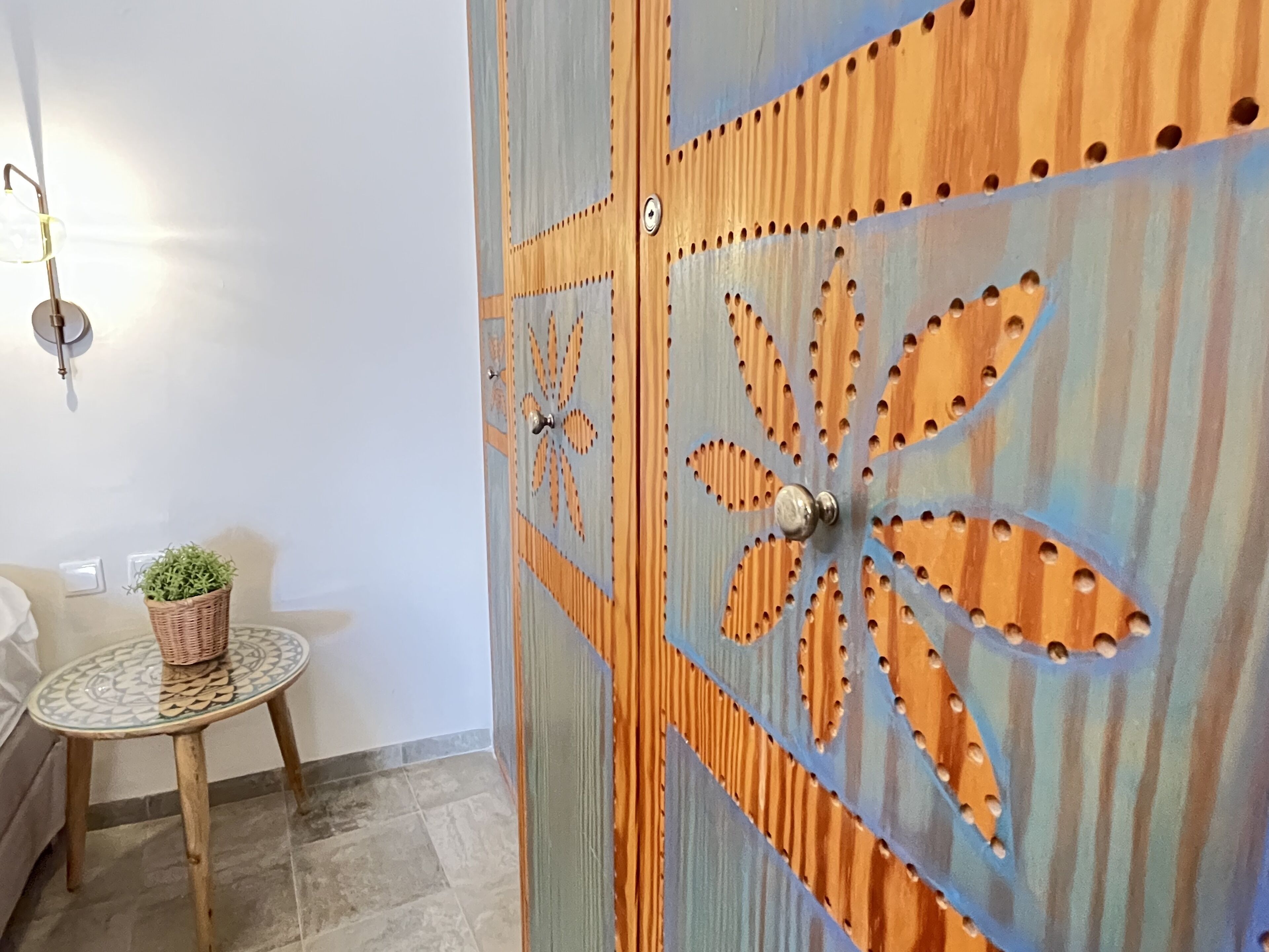 A room with a wooden door featuring a geometric pattern, a small round table with a woven basket, and a wall-mounted light fixture.