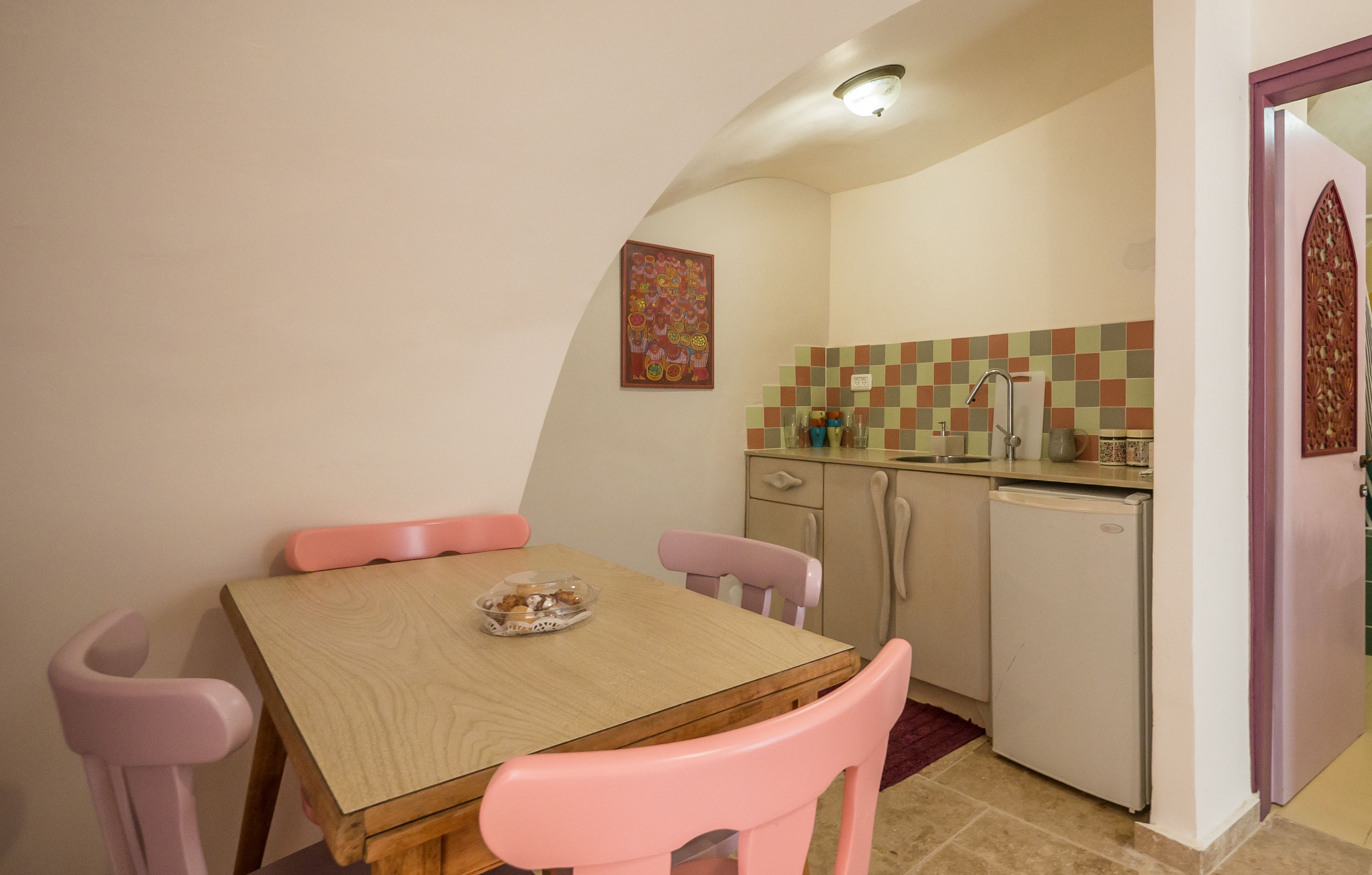 A compact kitchen with a wooden table and pink chairs, a small refrigerator, and a tiled backsplash.