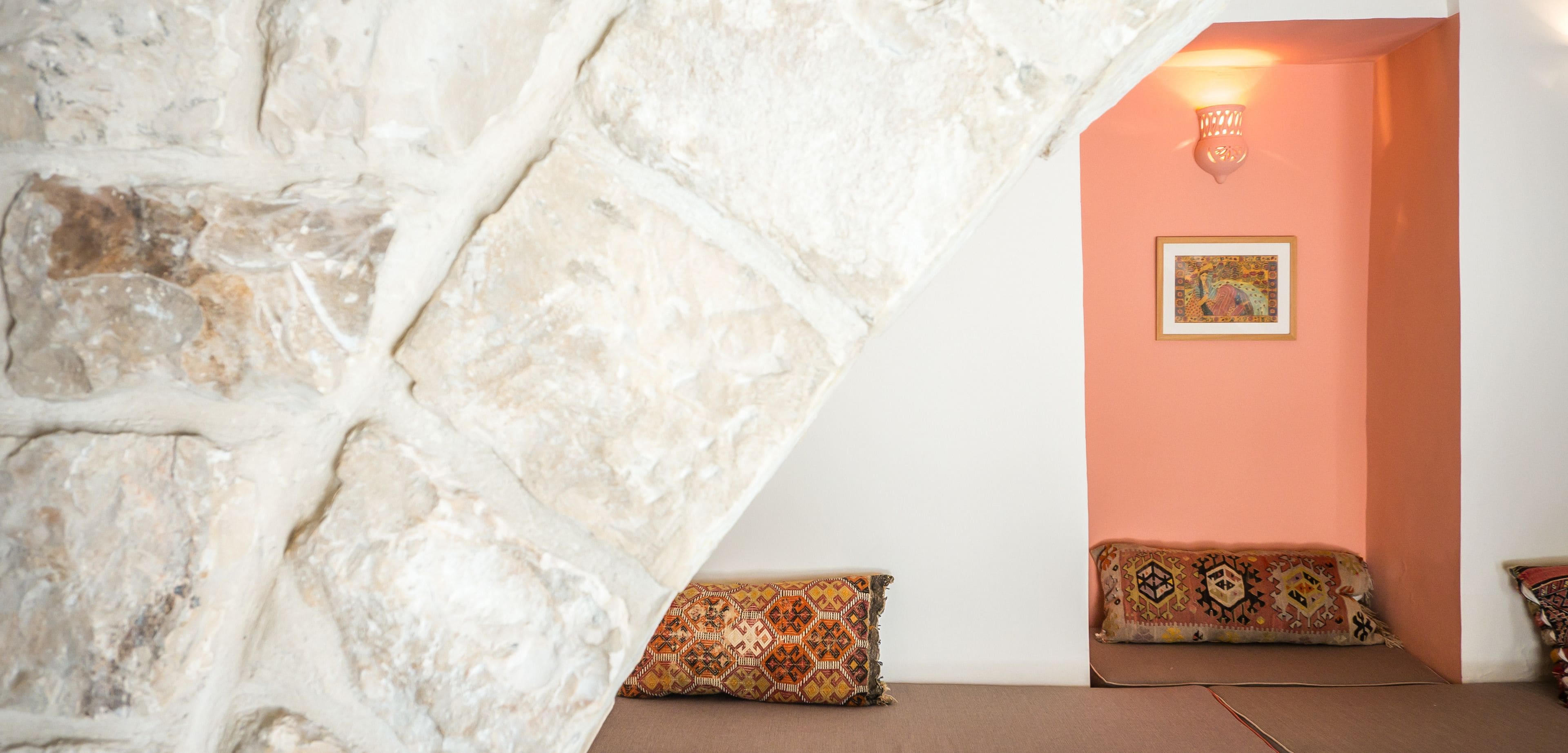 A room with a stone wall, a peach-colored wall, and a framed picture. There are two patterned cushions on a brown sofa.