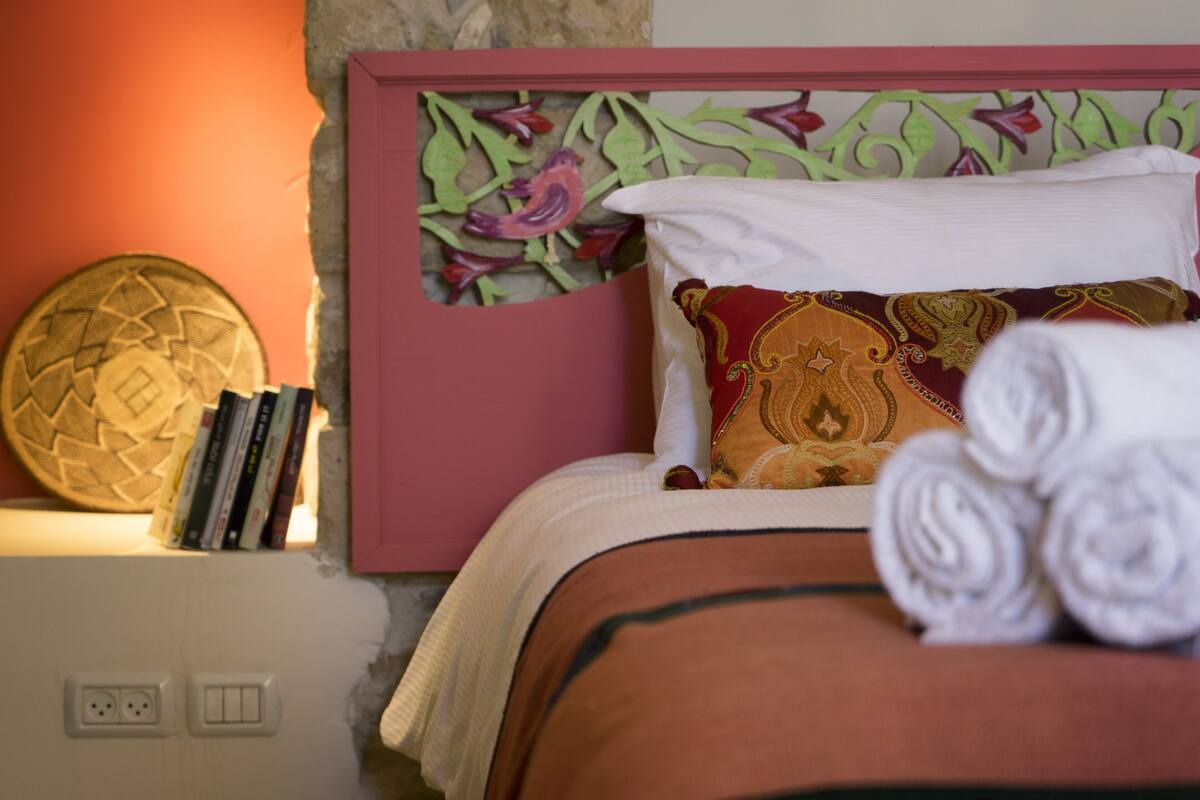 A neatly made bed with a patterned pillow and a rolled towel, a decorative wall piece, and a small shelf with books and a decorative item.