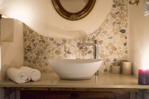 Un baño con un respingo de mosaico de piedra, un lavabo blanco tipo cuenco, un espejo con marco dorado y un mueble de tocador de madera.