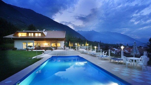 A swimming pool with lounge chairs and tables, a house with a terrace, and mountains in the background. - Pension Weihergut (Cermes)