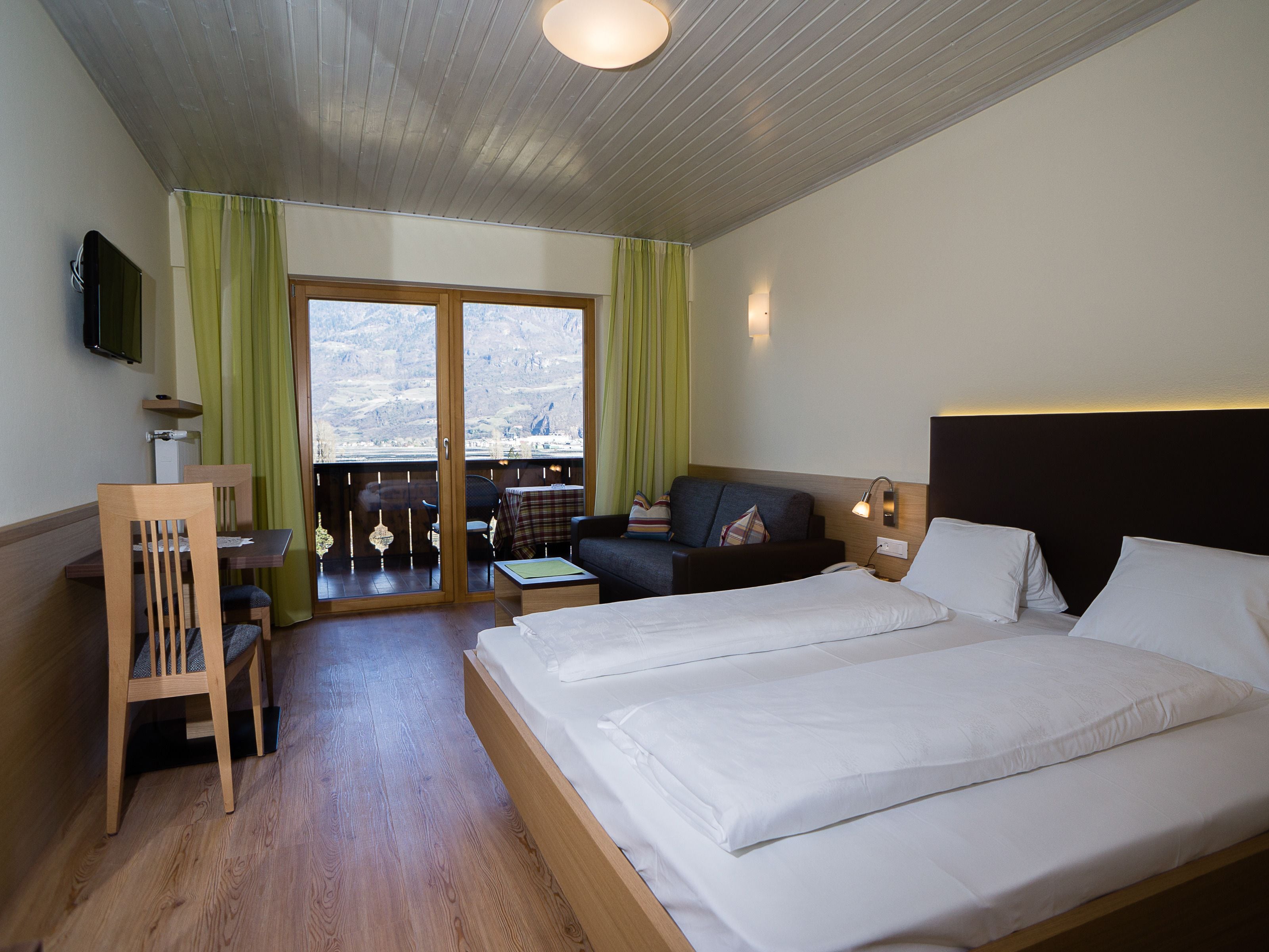 A hotel room with a bed, a desk, a chair, a sofa, and a view of the mountains.