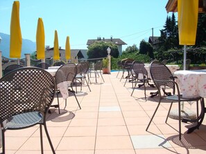 An outdoor patio area with metal chairs and tables, yellow umbrellas, and a view of houses and mountains in the background. - Pension Weihergut (Cermes)
