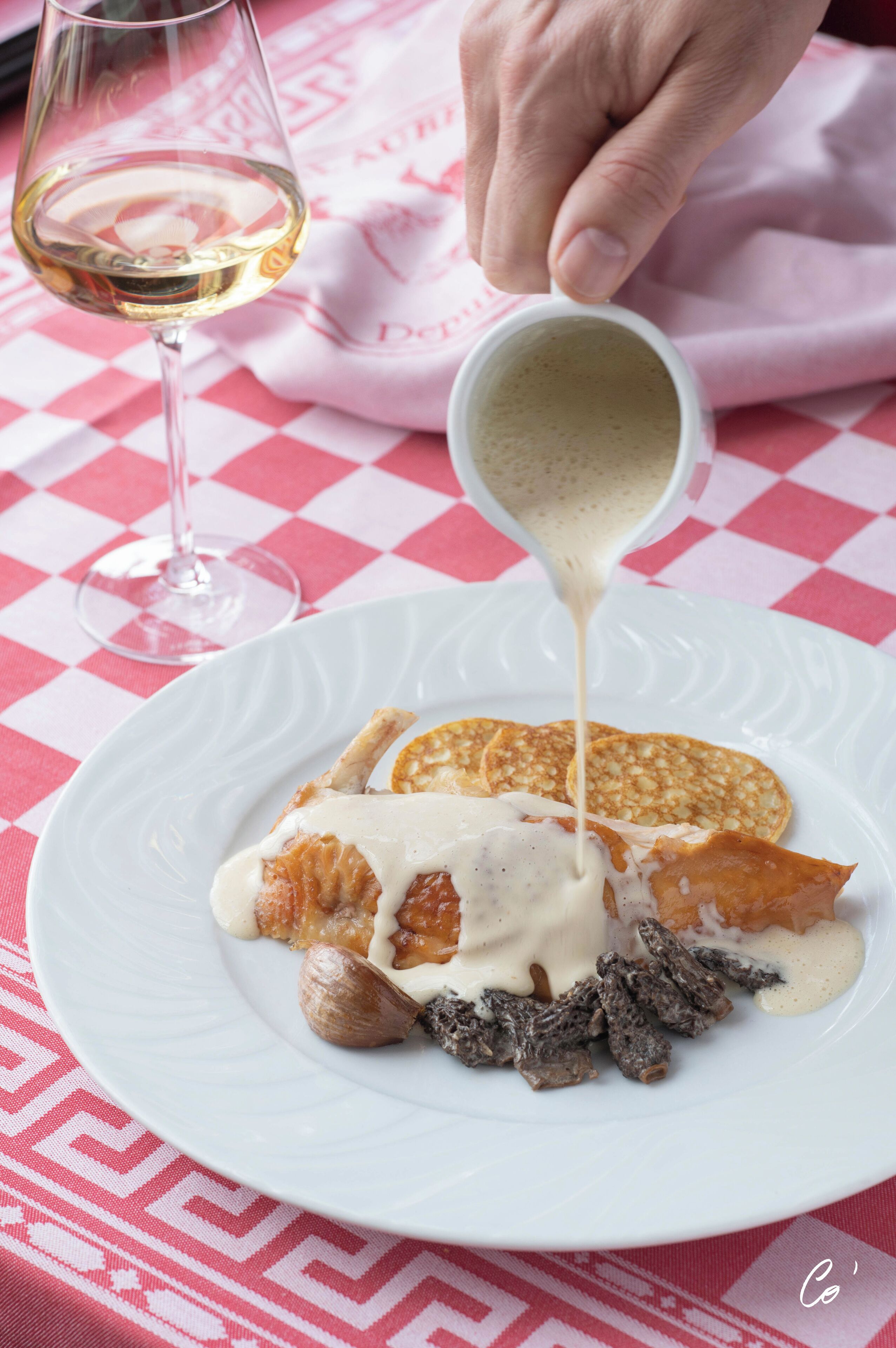 A white plate with pancakes, a glass of white wine, and a hand pouring sauce.