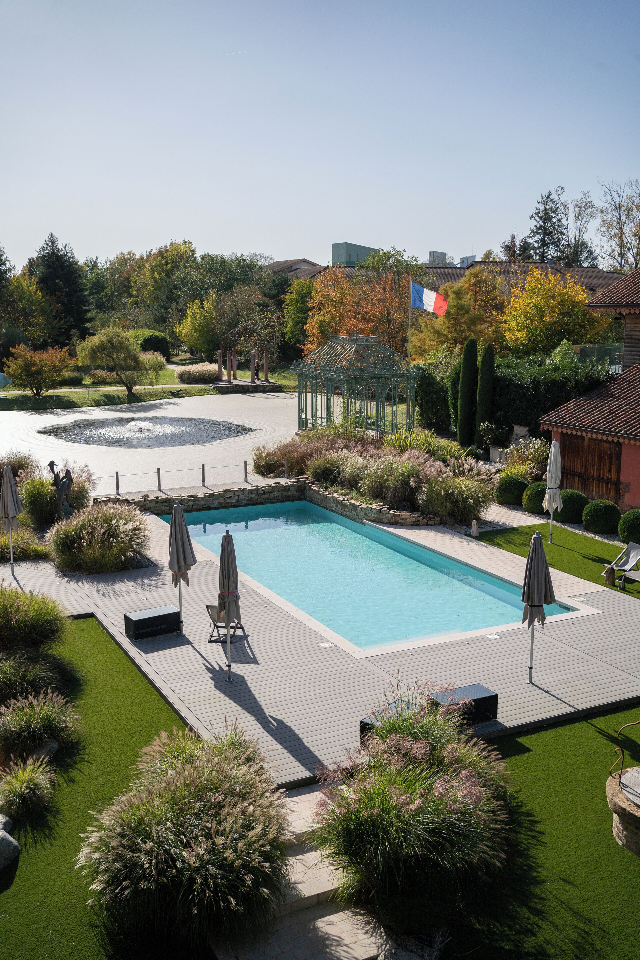A rectangular swimming pool with a surrounding deck, surrounded by well-maintained landscaping and a French flag.