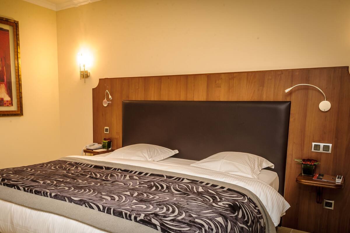 A hotel room with a large bed, a nightstand, a wall-mounted lamp, and a framed painting.