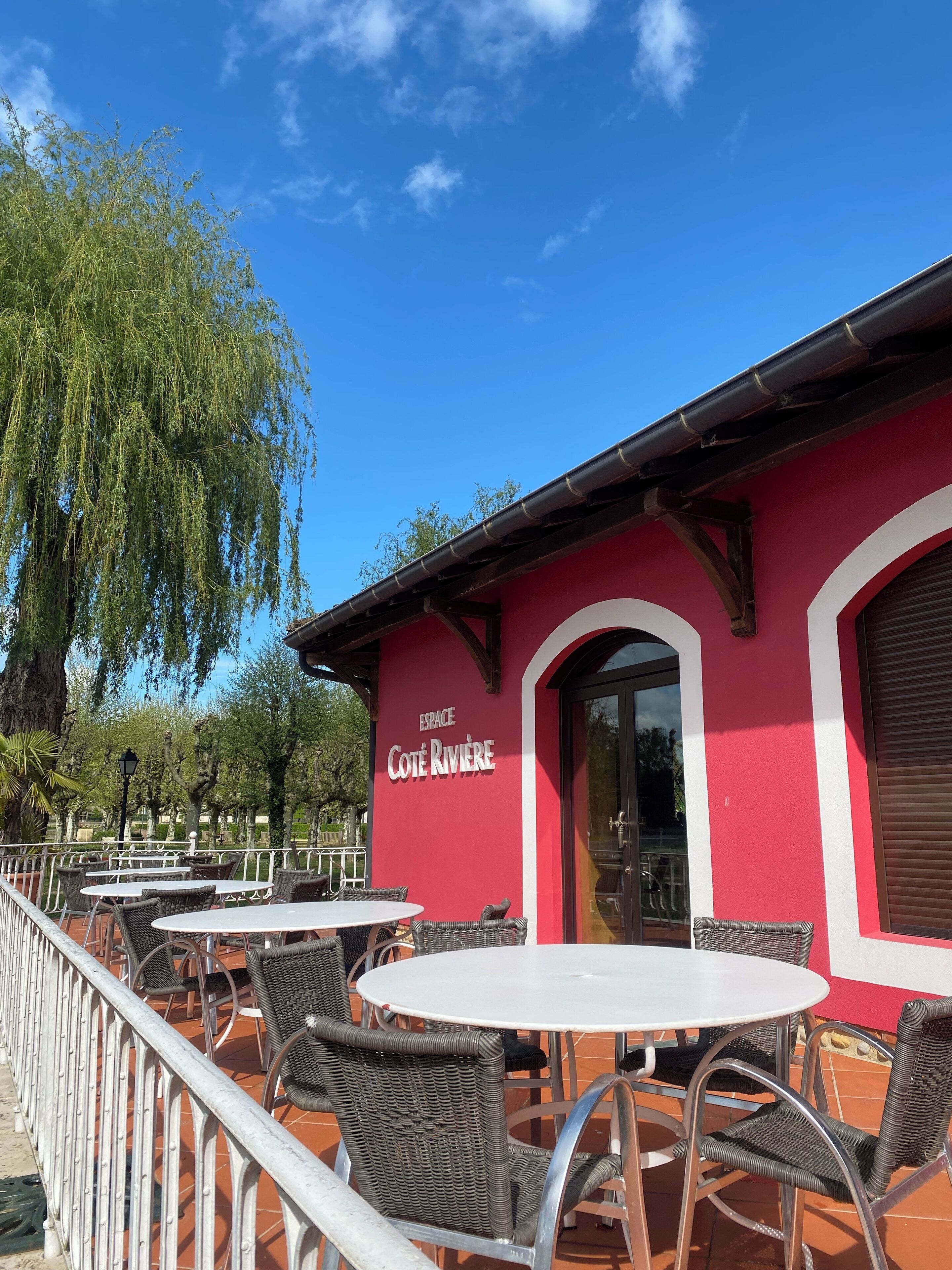 Un bâtiment rouge portant l’enseigne « ESPACE Côte Rivière » et doté d’une terrasse avec des chaises et des tables en osier.
