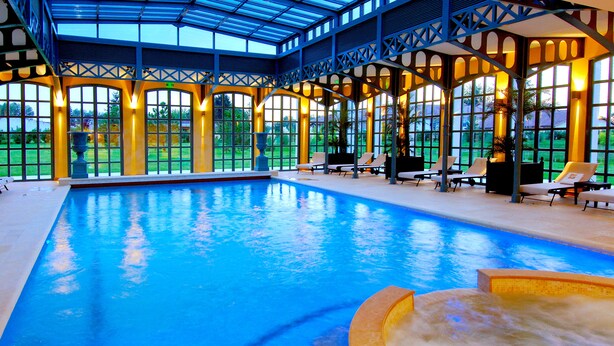 Una amplia piscina interior con techo de cristal, rodeada de sillones de descanso y vistas al verdor.