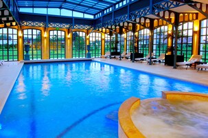 Una amplia piscina interior con techo de cristal, rodeada de sillones de descanso y vistas al verdor.