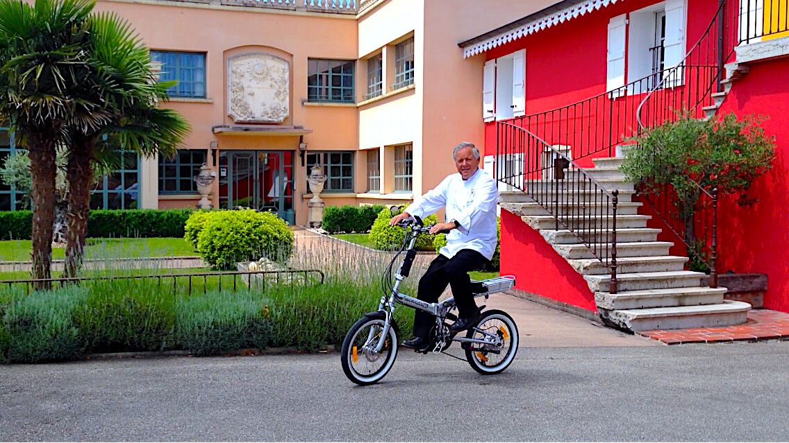 Un hombre pedalea en bicicleta frente a un edificio con fachada roja y una escalera.