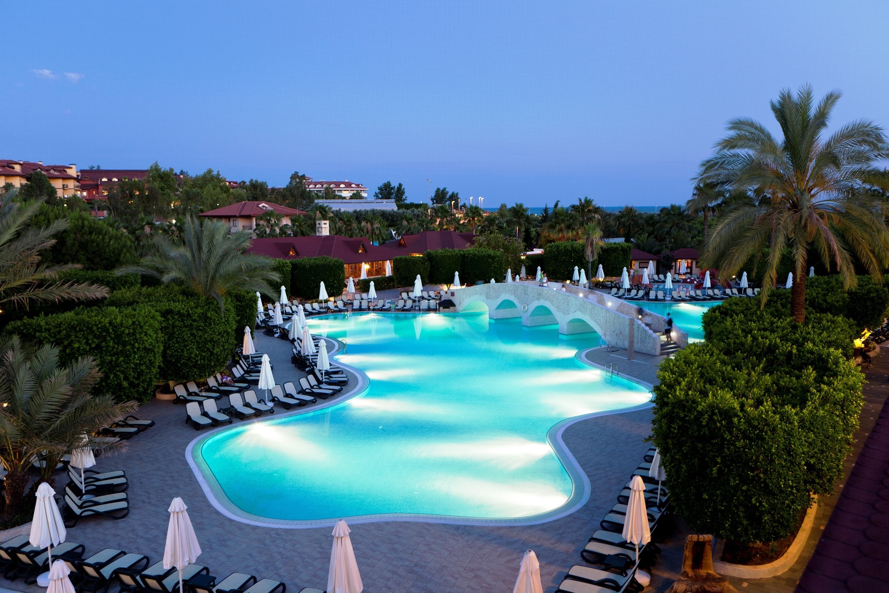 Une piscine d’hôtel avec des chaises longues et des parasols, entourée de palmiers et de bâtiments.