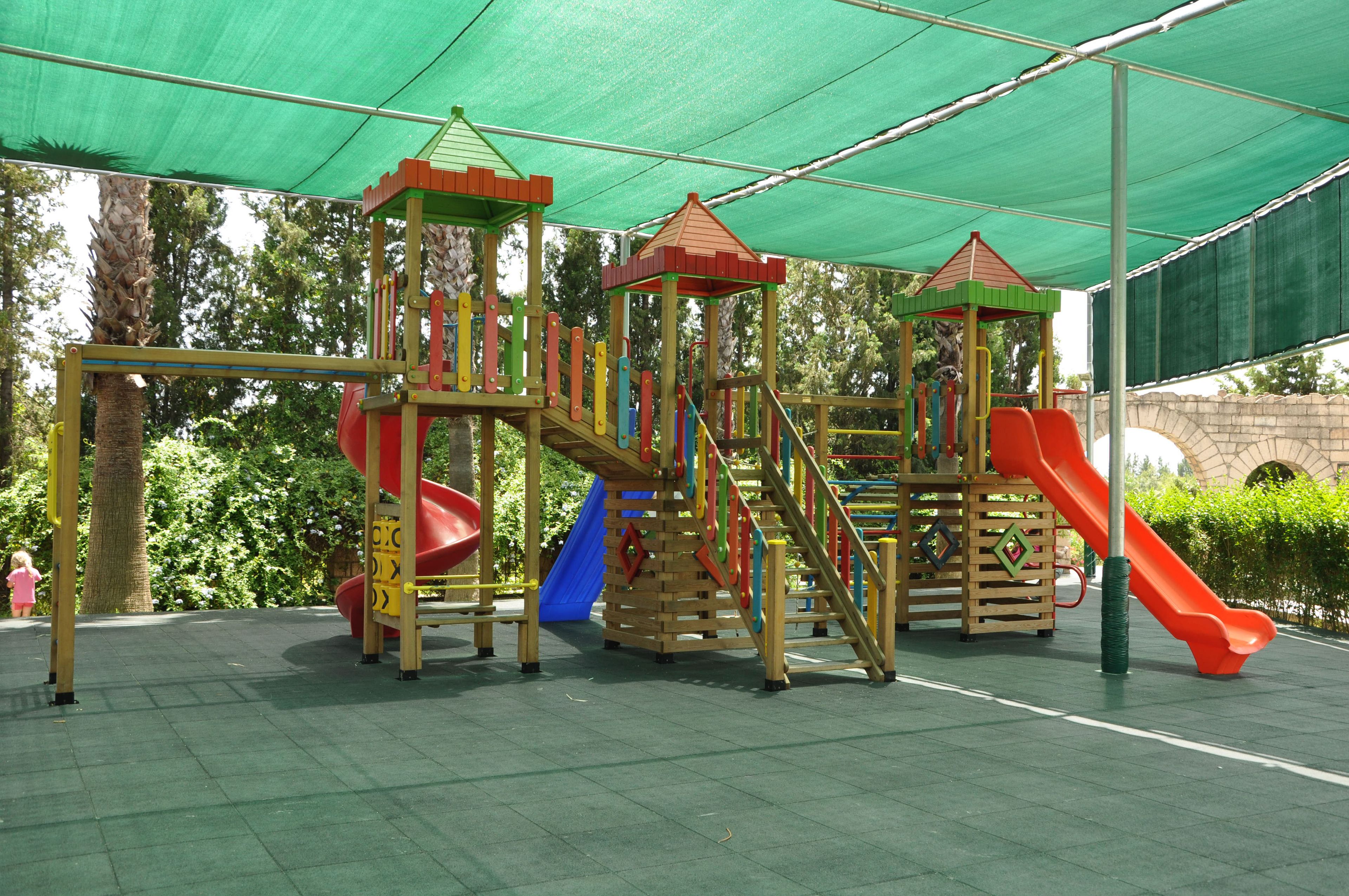 A playground with multiple slides and climbing structures, surrounded by a green canopy.
