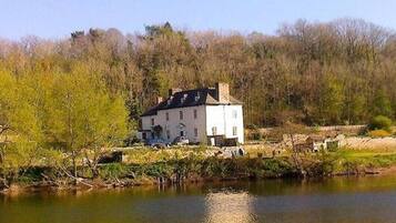 A white building with a black roof, situated by a body of water, surrounded by trees and greenery.