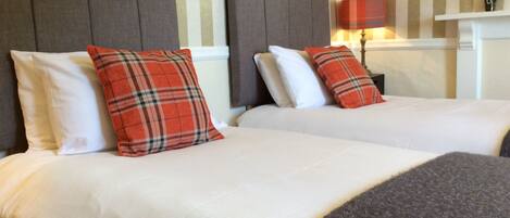 Une chambre d’hôtel avec deux lits, chacun agrémenté de draps blancs et d’oreillers à carreaux rouges. La chambre est décorée de papier peint à rayures et comprend une lampe sur une table de chevet.