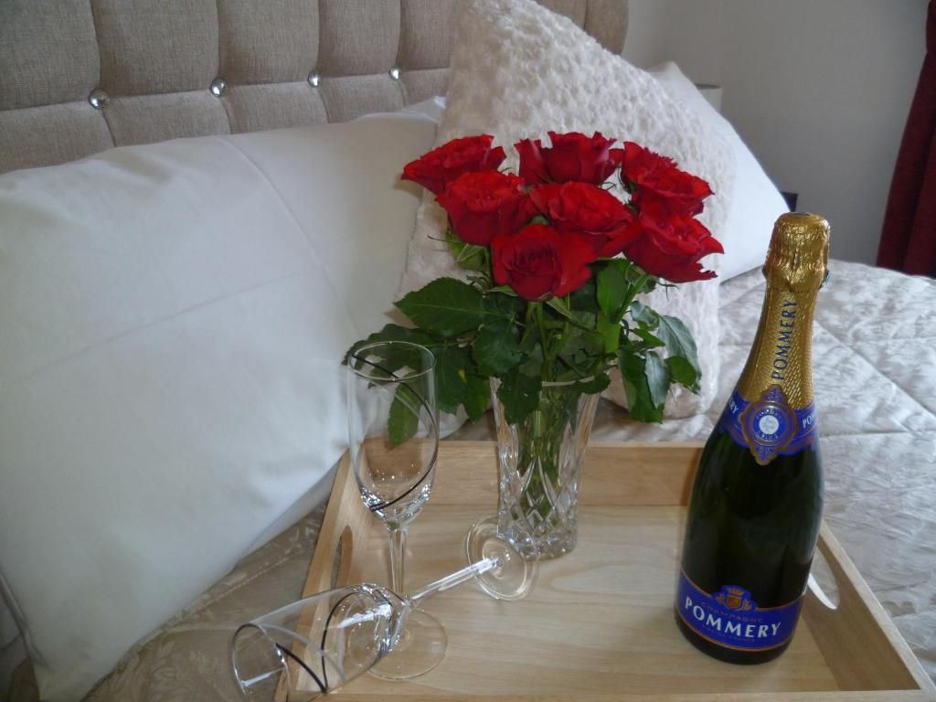 Ein Bett mit einer gepolsterten Kopfstütze, ein Nachttisch mit einer Champagnerflasche, eine Vase mit roten Rosen und zwei Weingläser.