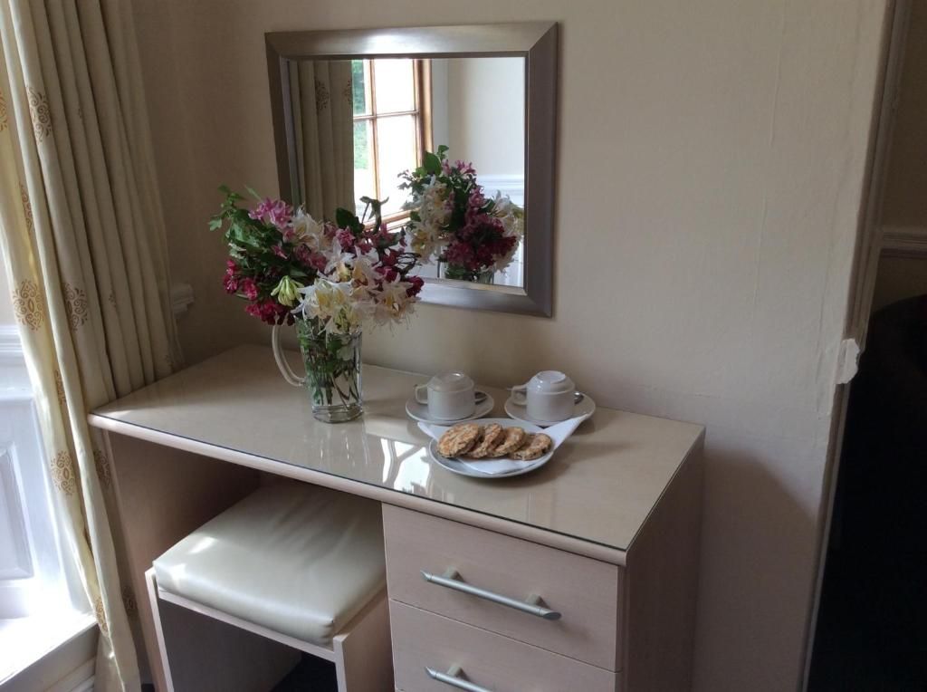 A room with a vanity table, a mirror, a vase with flowers, two cups, and a plate with cookies.