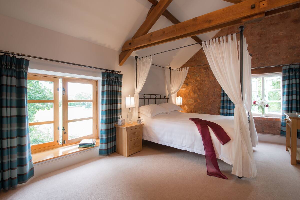 Quarto com cama de dossel, mesa de cabeceira, janela com cortinas e parede de pedra.
