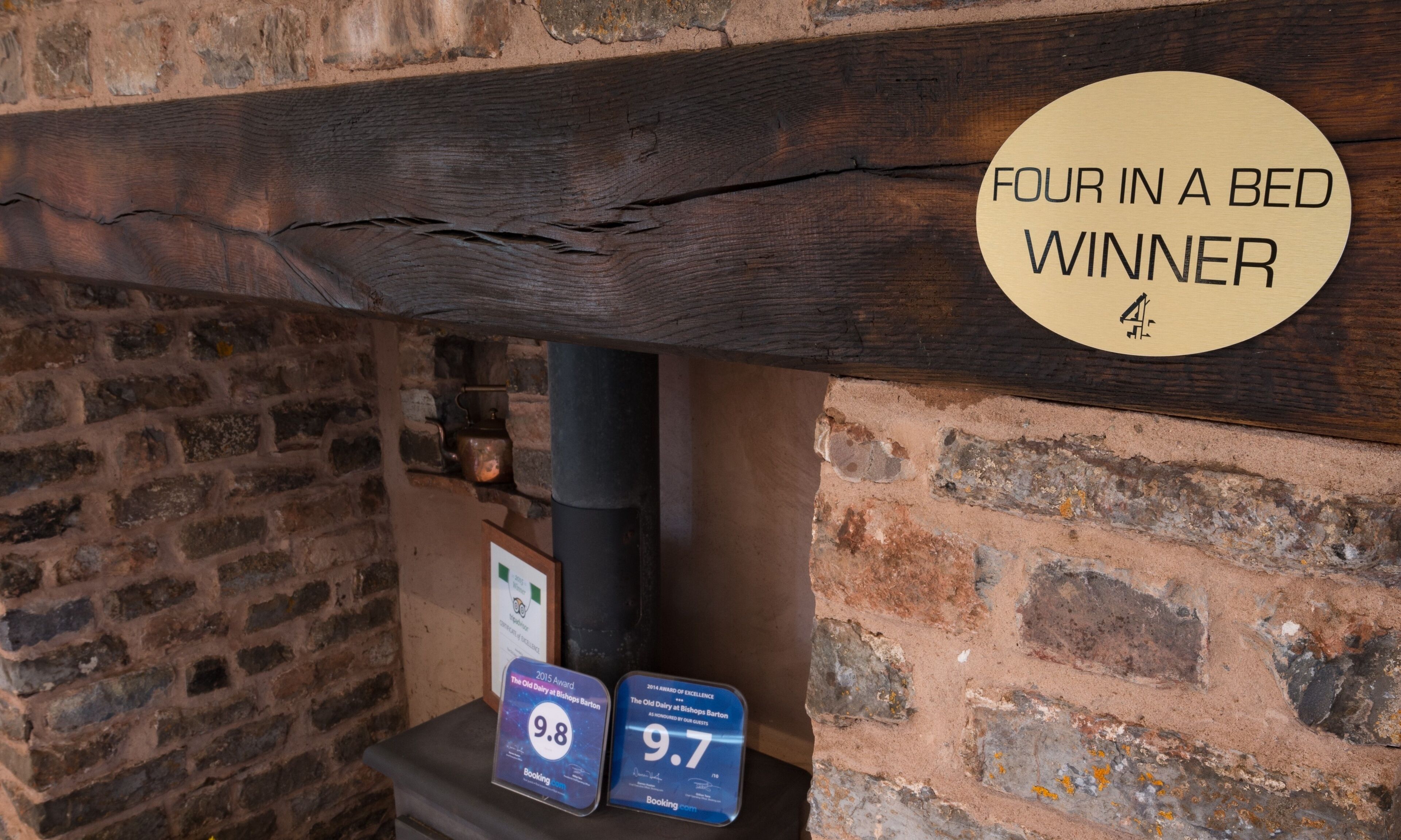 Una pared de ladrillo rústica con una viga de madera sobre una chimenea. Hay un cartel que indica 'Four in a Bed Winner' y dos premios con valoraciones de 9.8 y 9.7.