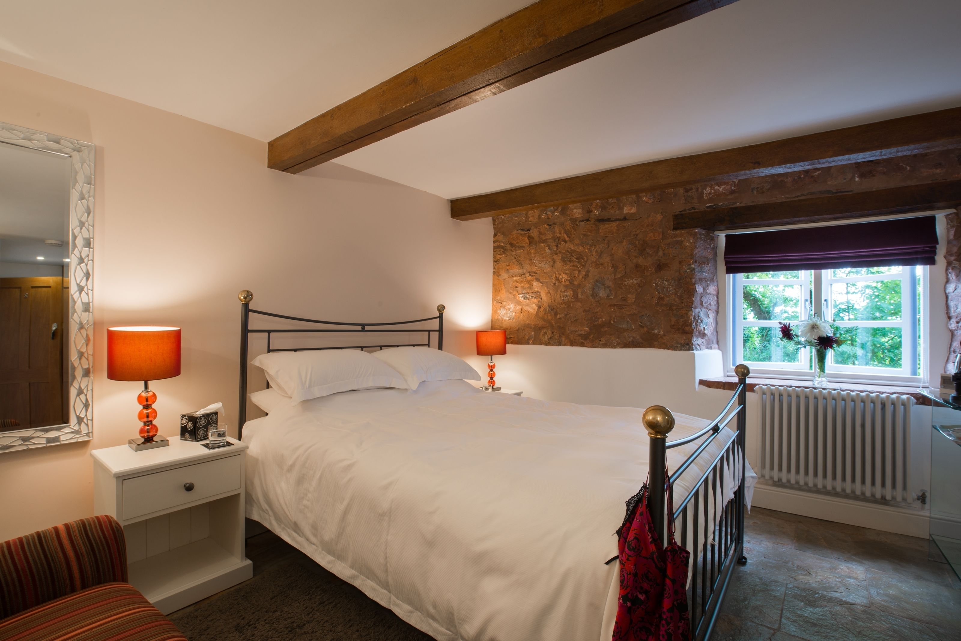 A bedroom with a large bed, two bedside tables with lamps, a window with a view, and a stone wall.