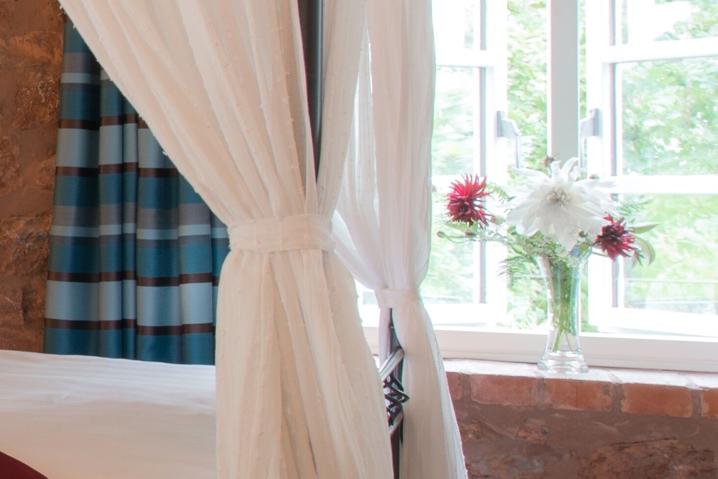 A room with a bed, a window with curtains, and a vase with flowers on the windowsill.