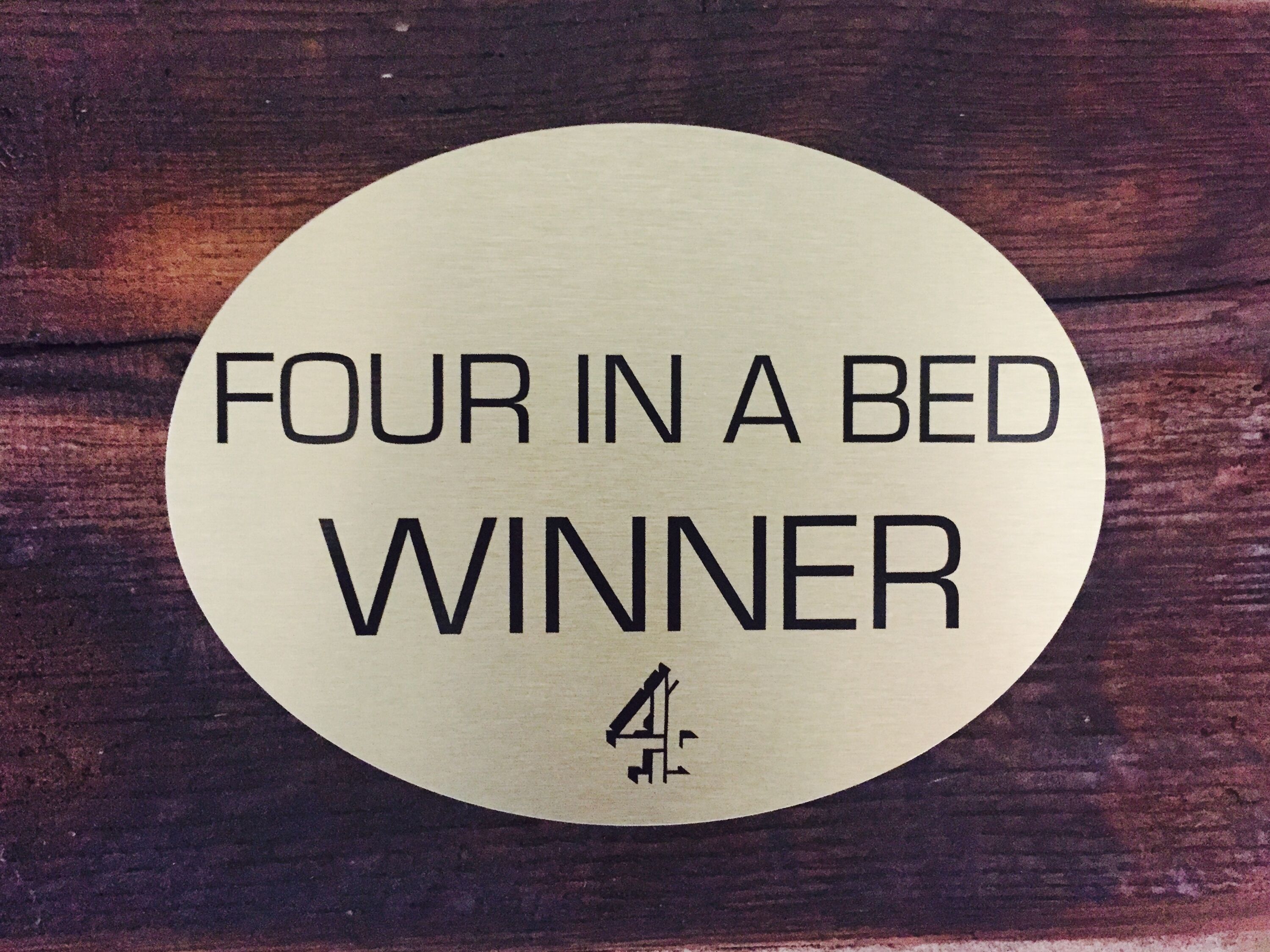 A circular sign with the text "FOUR IN A BED WINNER" and the number "4" below it, placed on a wooden surface.