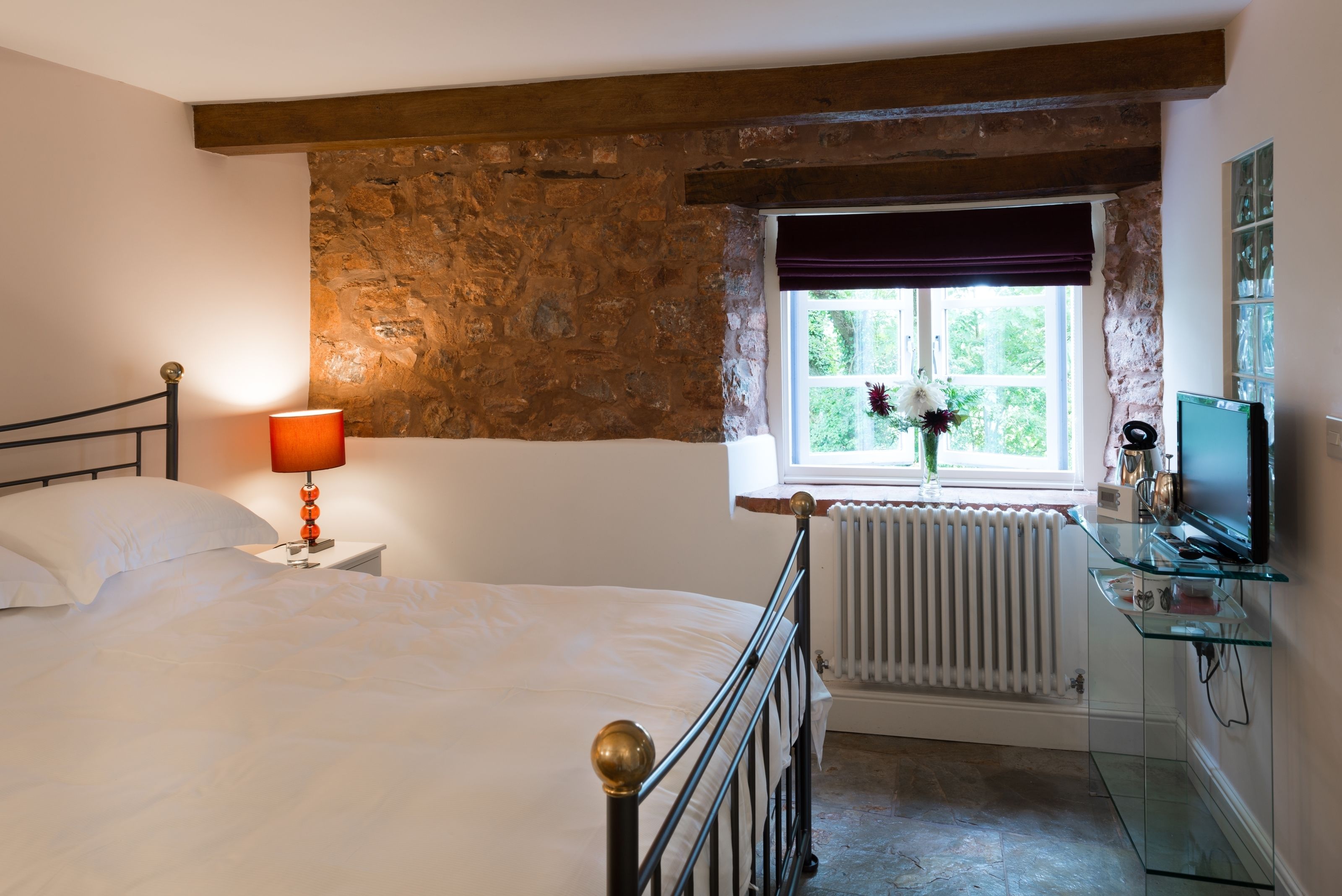 A bedroom with a stone wall, a bed with white linens, a bedside lamp, a radiator, a window with a view, and a glass TV stand.