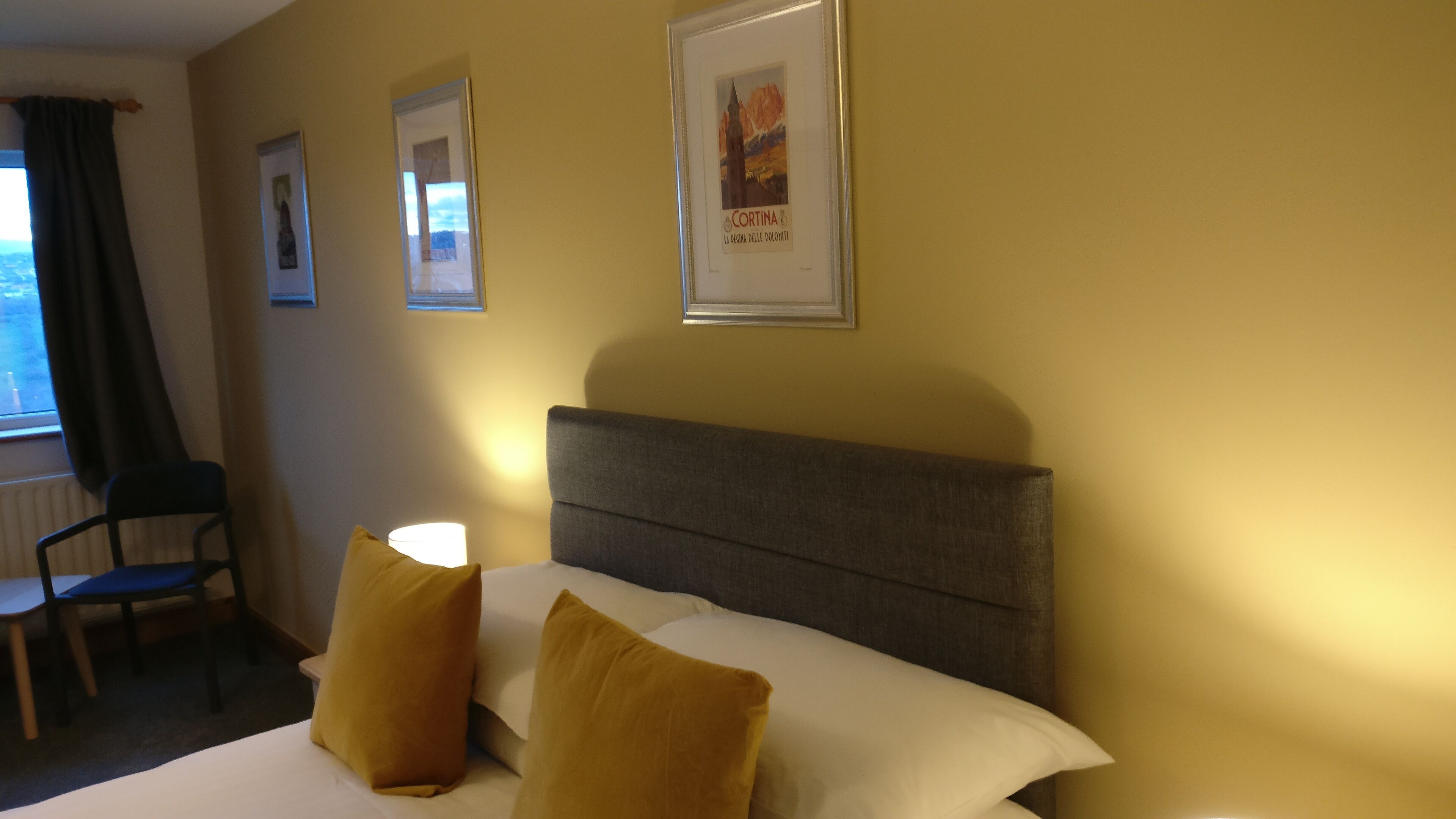 A hotel room with a bed, two pillows, a chair, a lamp, and a framed picture on the wall.