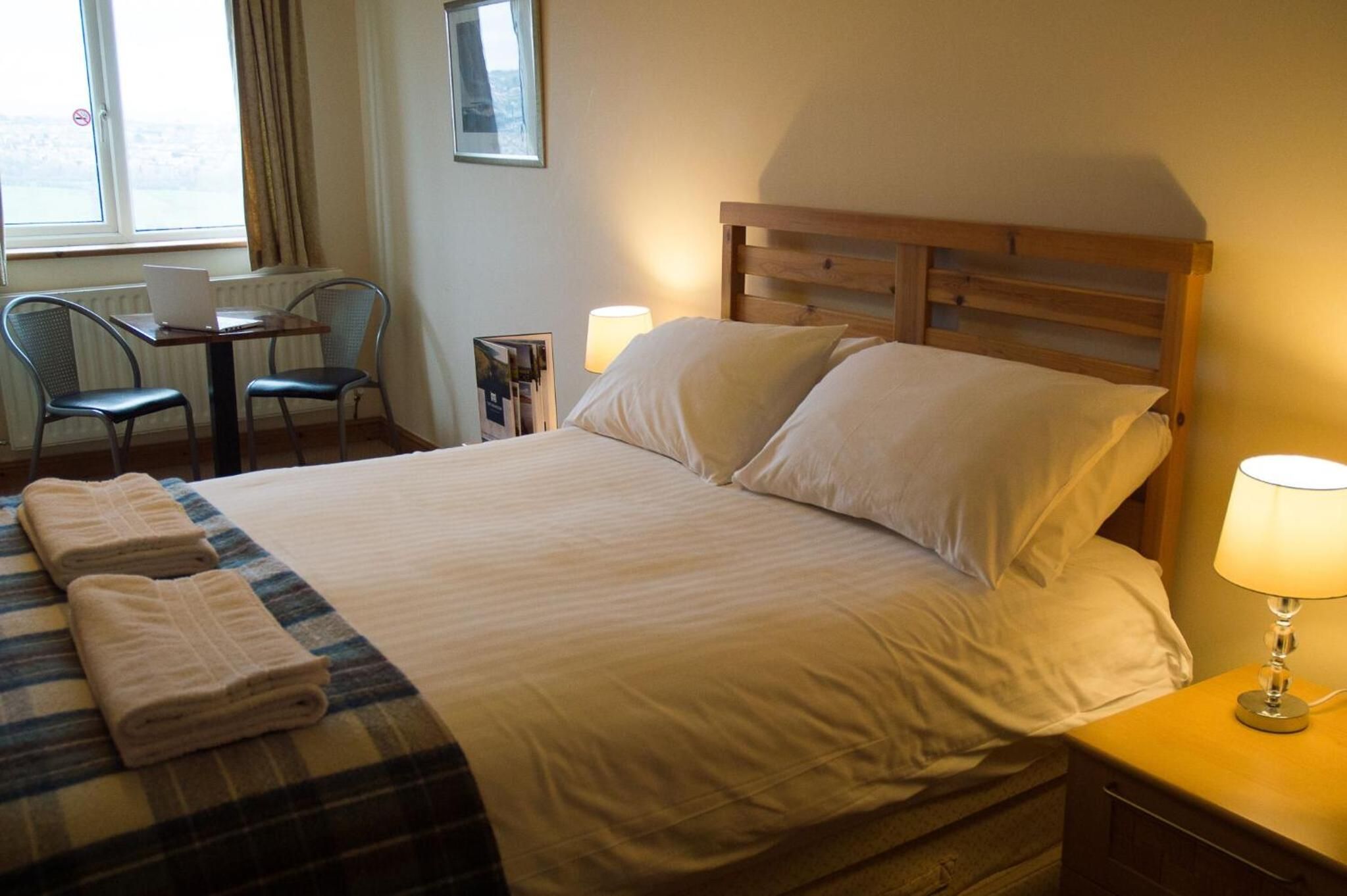 Une chambre d’hôtel avec un lit, deux lampes de chevet, une chaise, une table avec un ordinateur portable et une fenêtre avec des rideaux.