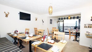A dining area with multiple tables set for meals, a television mounted on the wall, and a view of the sea through the windows. - Ardentorrie Guest House (Inverness)