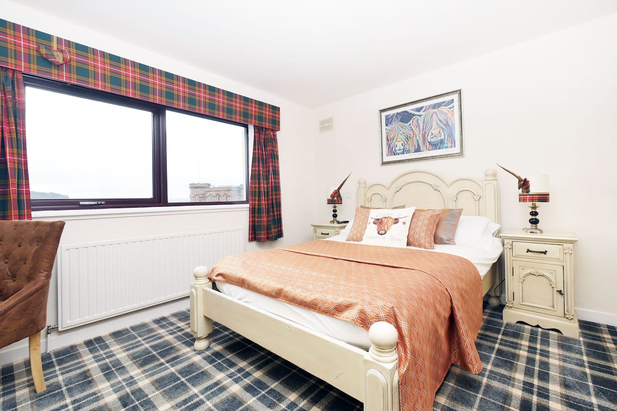 A bedroom with a bed, a chair, a window with plaid curtains, a radiator, a nightstand with a lamp, and a framed picture on the wall.