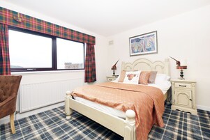A bedroom with a bed, a chair, a window with plaid curtains, a radiator, a nightstand with a lamp, and a framed picture on the wall.