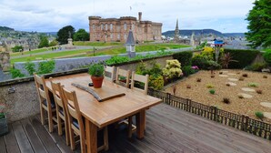 A wooden deck with a table and chairs, overlooking a landscaped garden and a historic building in the background. - Ardentorrie Guest House (Inverness)
