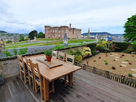 Una terraza de madera con una mesa y sillas, con vistas a un jardín ajardinado y un edificio histórico al fondo.