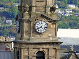 Una torre de reloj con un gran reloj, arquitectura de piedra y vistas de casas al fondo.